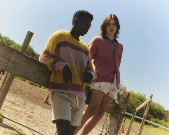 Two young models leaning against a wooden fence outdoors under a clear blue sky, wearing colorful knit sweaters.