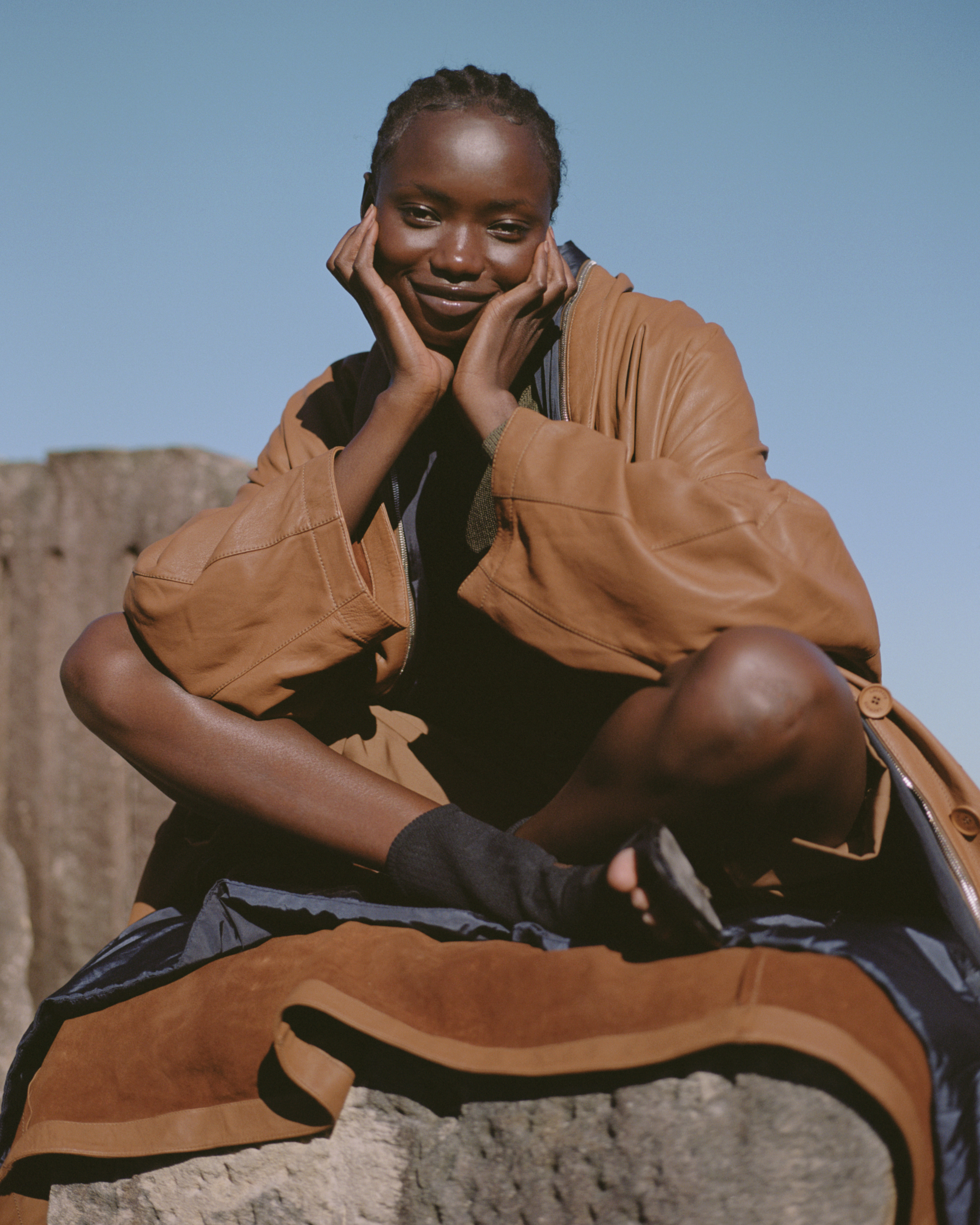 Woman in a brown coat sits cross-legged on a rock formation under a blue sky, smiling with her hands on her cheeks.