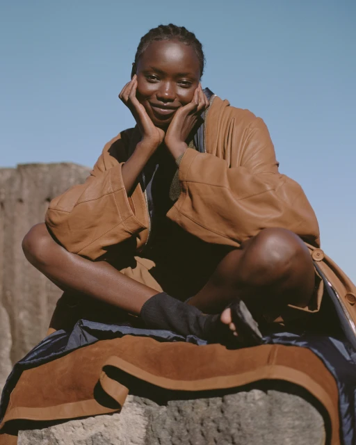 Woman in a brown coat sits cross-legged on a rock formation under a blue sky, smiling with her hands on her cheeks.