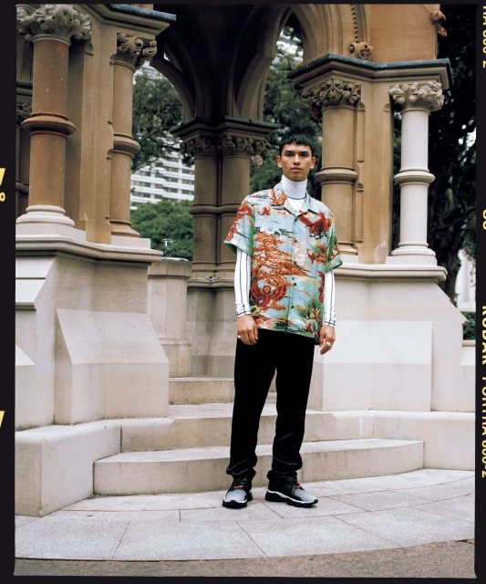 Man in patterned shirt and black pants stands in front of stone structure in urban park setting, medium shot, neutral expression.
