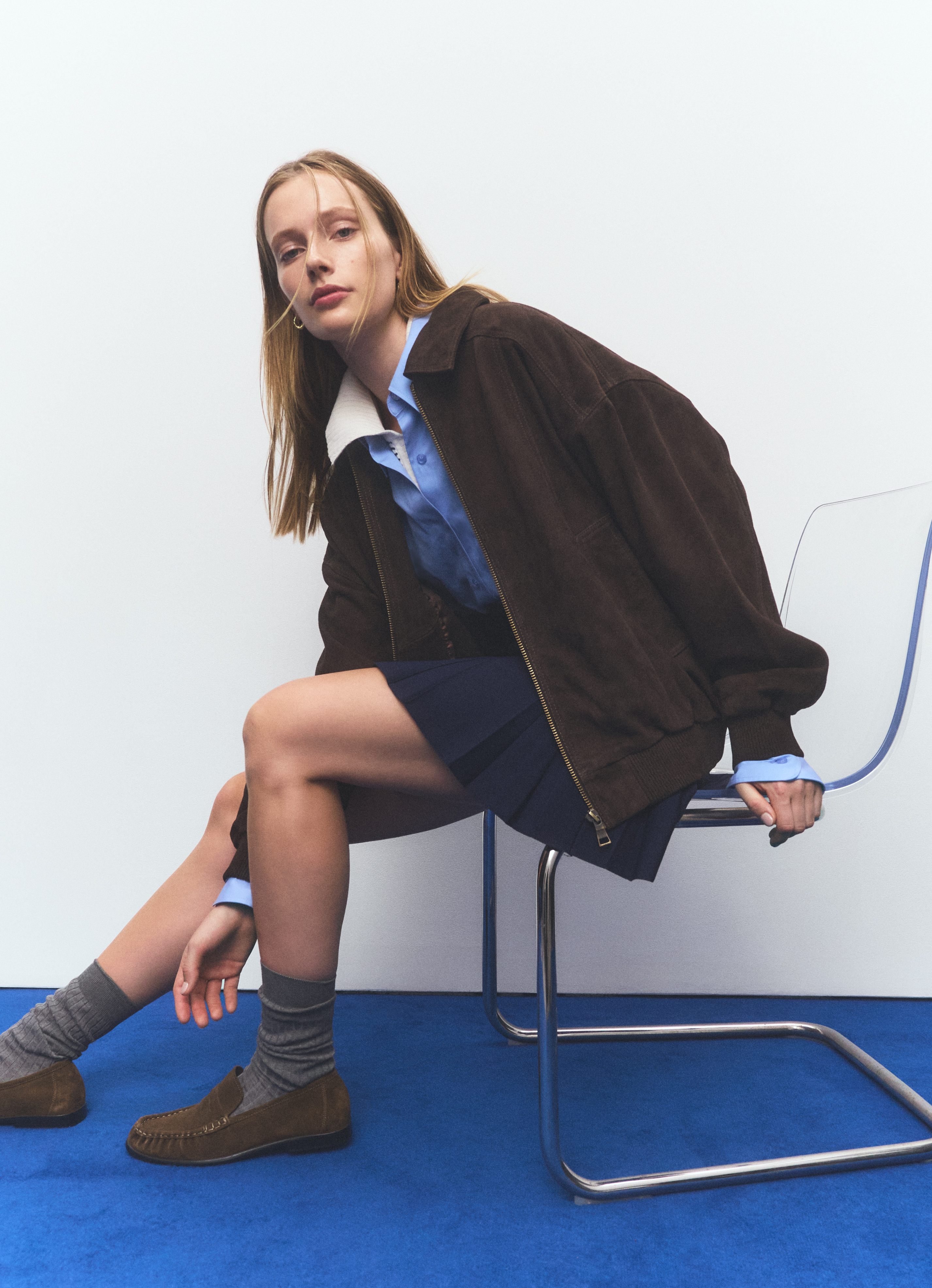 A fair-skinned woman sits on a clear chair wearing a brown bomber jacket, blue shirt, navy skirt, grey socks, and brown moccasins.