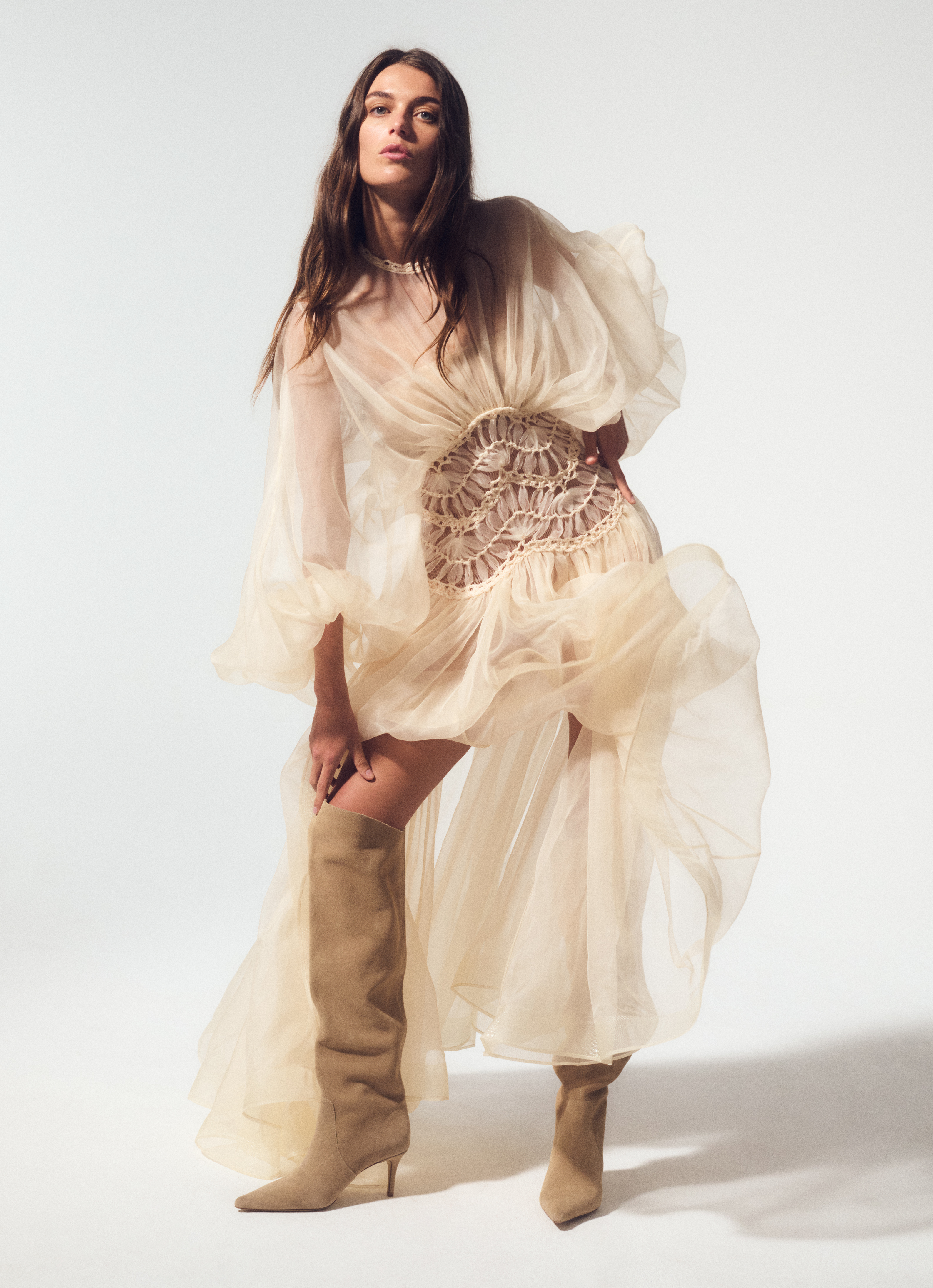 A woman poses in a sheer, ruffled dress and knee-high boots against a white background in a fashion forward portrait.
