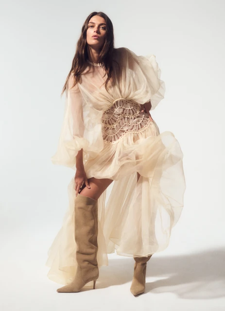 A woman poses in a sheer, ruffled dress and knee-high boots against a white background in a fashion forward portrait.