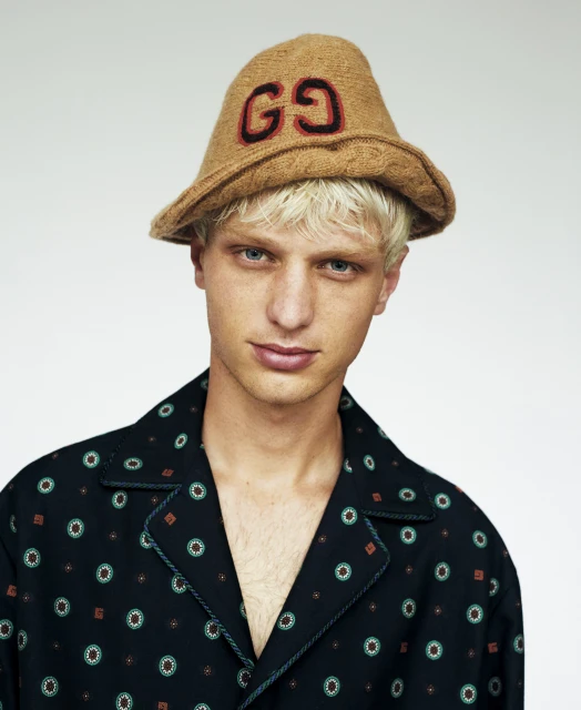 Portrait of a young adult male wearing a tan bucket hat with a 'GG' logo, posing against a neutral background in a patterned pajama top.