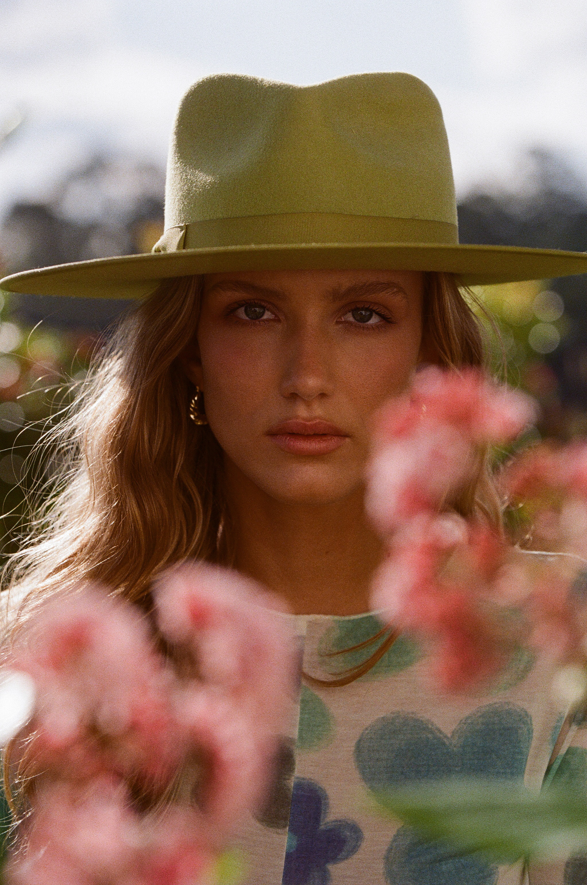 Portrait of a woman with wavy hair wearing a light green hat, adorned with earrings, standing outdoors amidst flowers and greenery.