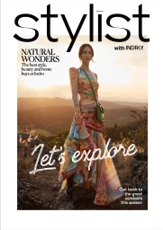 Model standing on a rocky mountain ridge at sunset wearing a colourful patterned one-shoulder dress and holding a green crossbody bag.