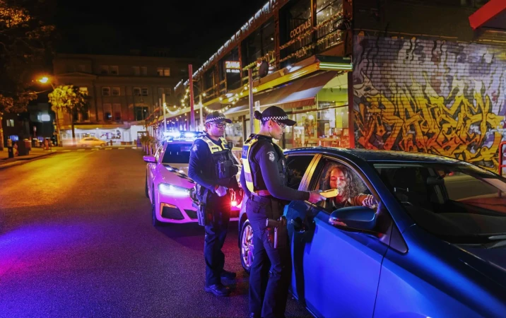 Two police officers conduct a roadside breath test on a woman seated in her car on a city street at night with ambient lighting.