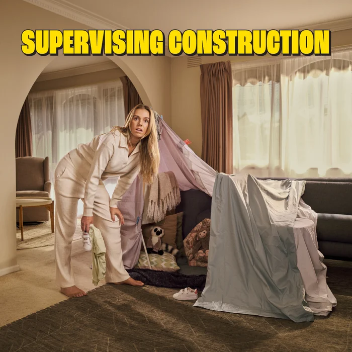 Woman in cream lounge set standing near a blanket fort in a living room holding baby shoes