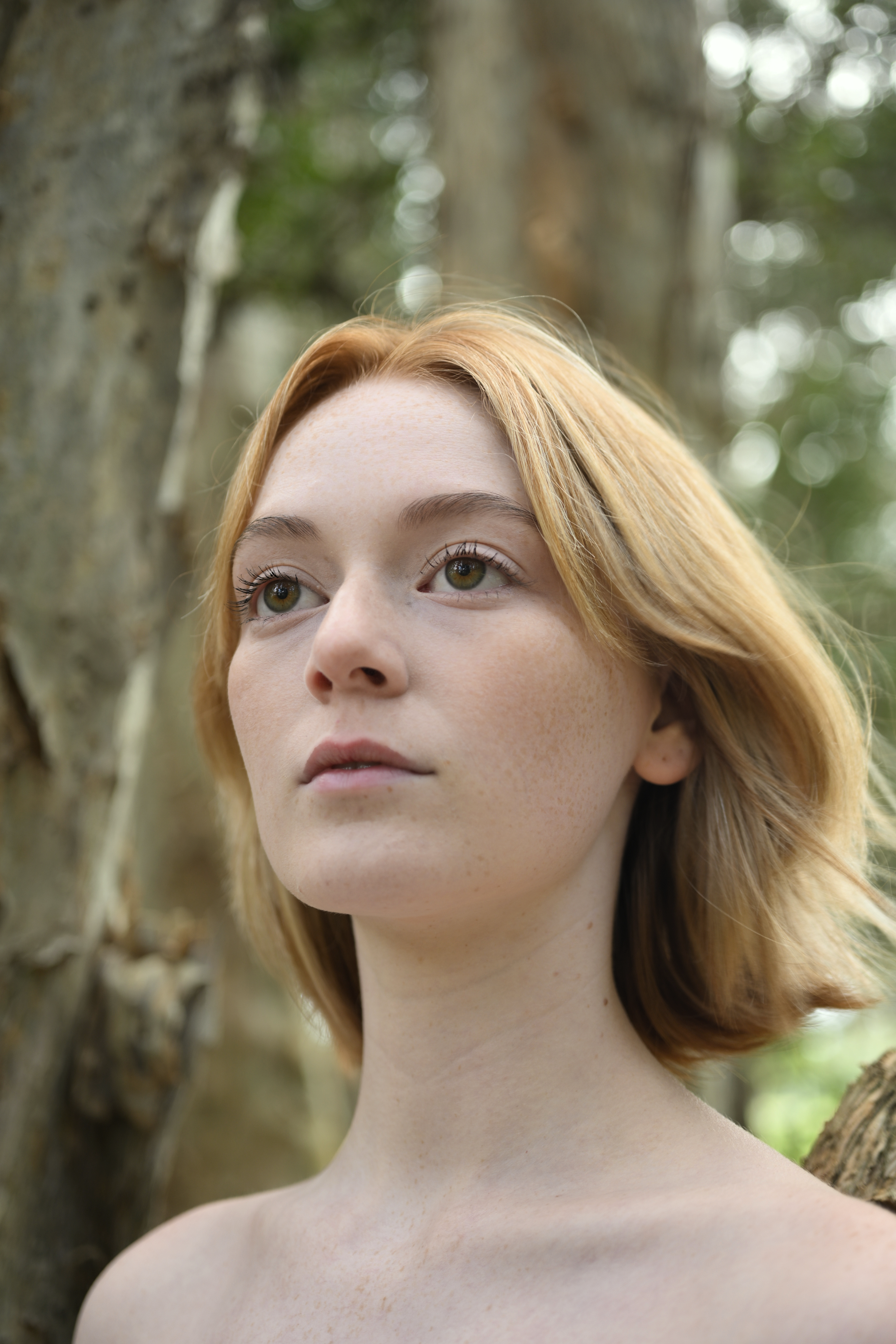 Portrait of a young adult female with fair skin and blonde hair, looking off-camera in an outdoor, wooded environment with soft lighting.