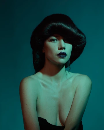 Glamour shot of a woman with dark hair and dark lipstick posing against a teal background in a beauty or fashion context.