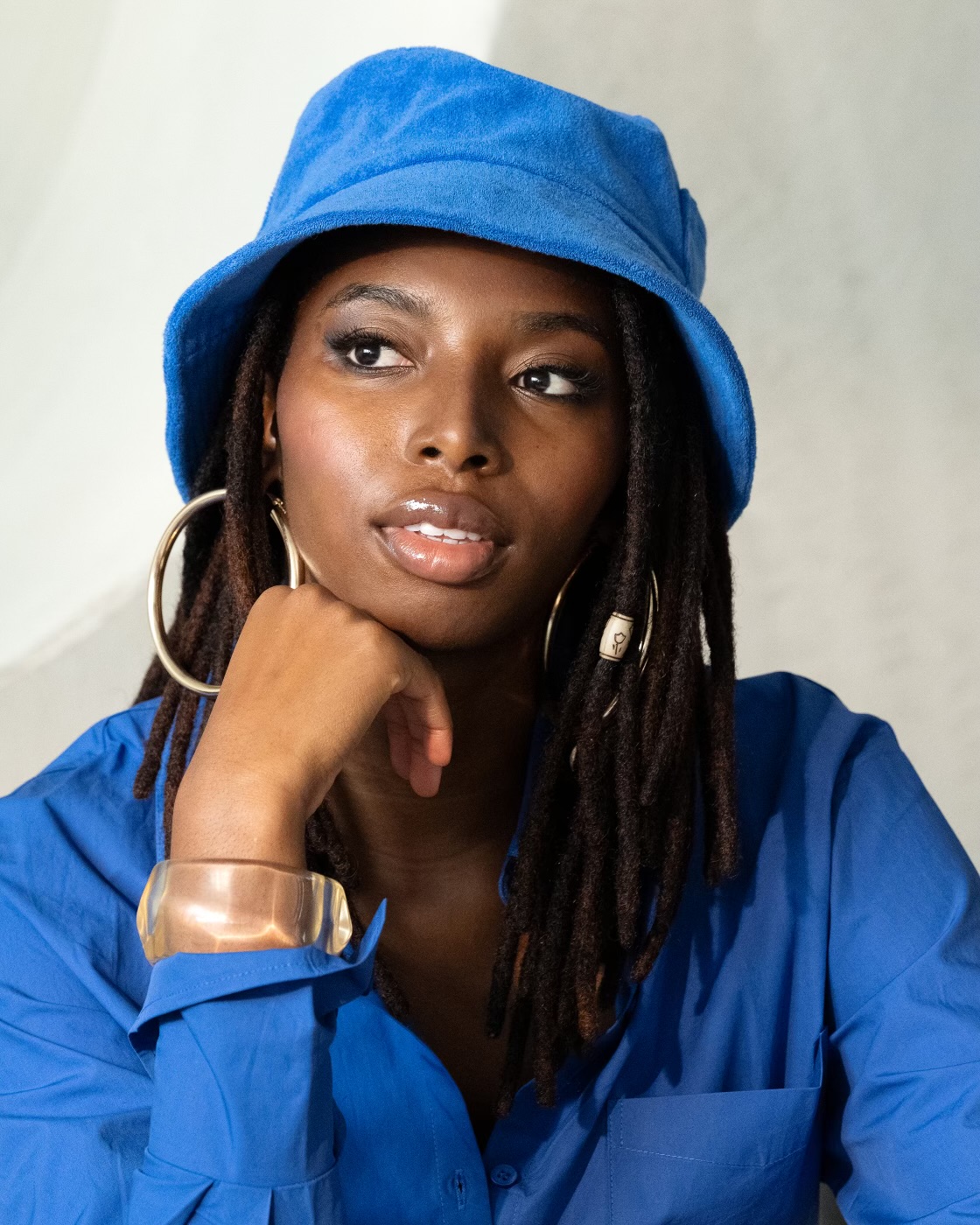 A black woman is posing in a photo studio wearing a bright blue bucket hat and a matching shirt, complemented by gold hoop earrings.