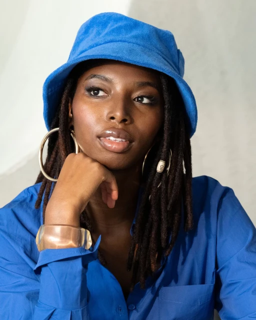 A black woman is posing in a photo studio wearing a bright blue bucket hat and a matching shirt, complemented by gold hoop earrings.