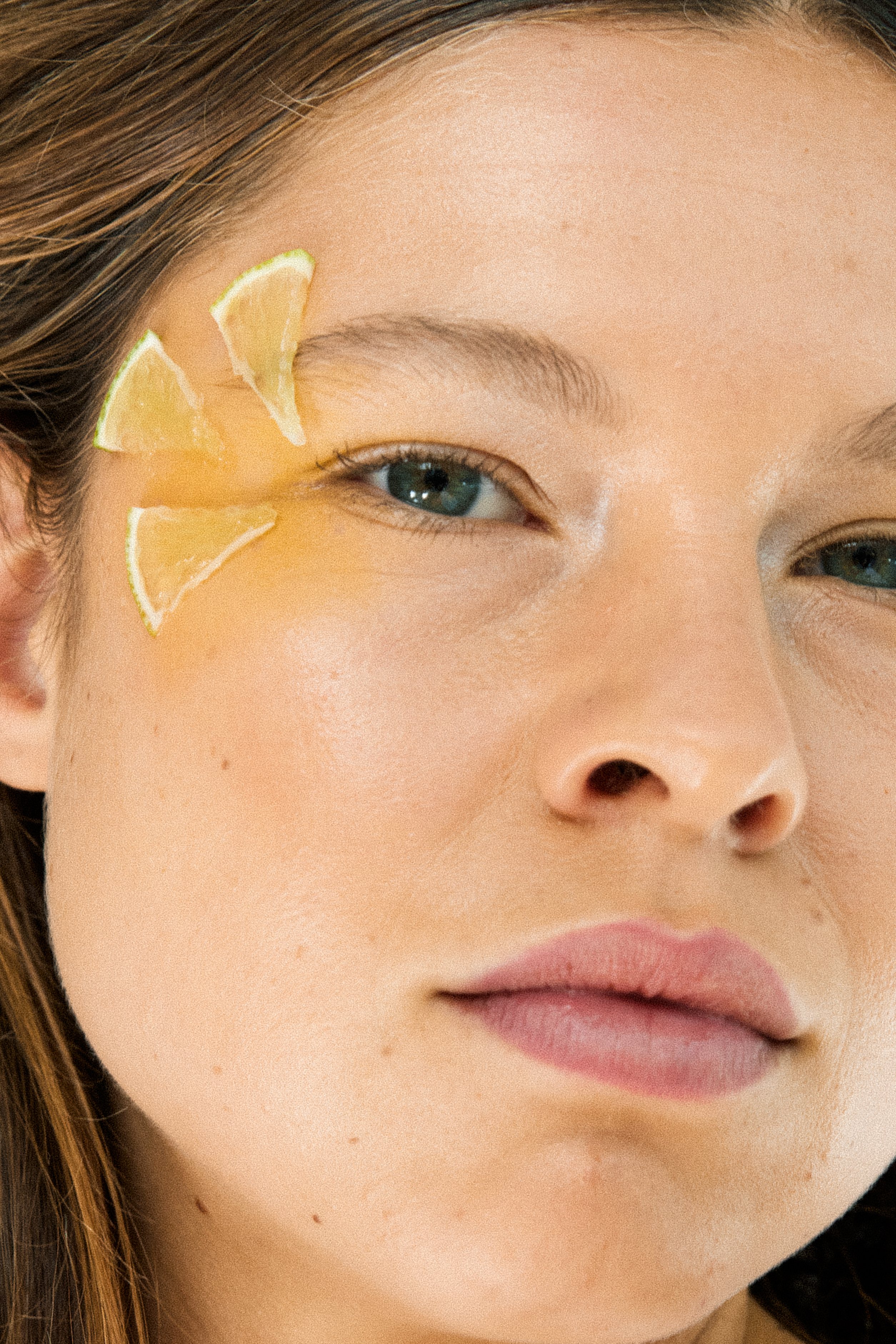 Close-up of a woman's face with three lemon wedges placed artfully on her forehead and cheek in a beauty or wellness context.