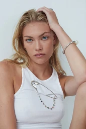 Close-up portrait of a blonde woman wearing a white tank top with a large silver safety pin brooch and a pearl bracelet.