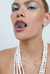 Close-up portrait of a woman with metallic silver eyeshadow holding an ice cube on her tongue and wearing a white shell necklace.