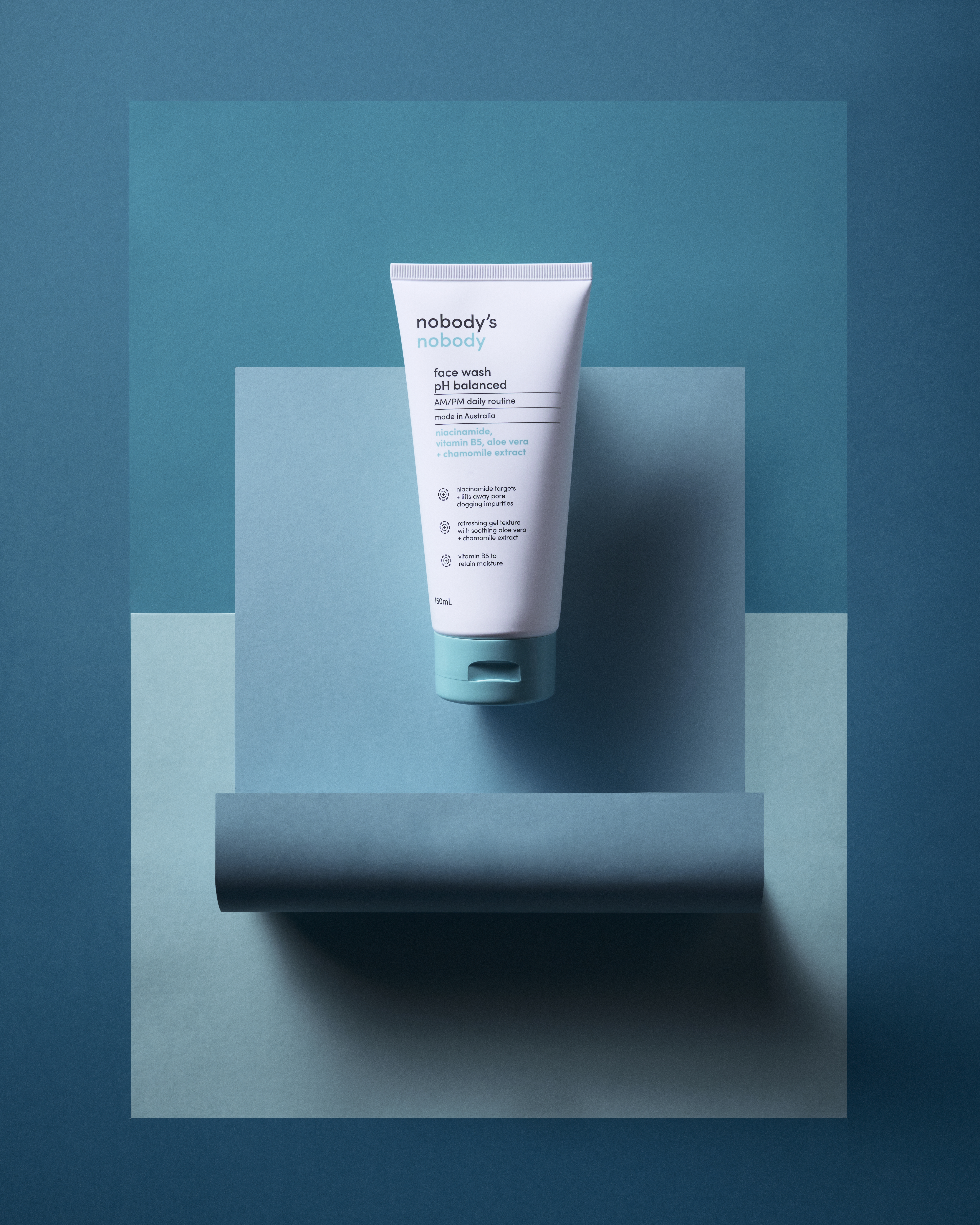Studio shot of a white tube of face wash on a teal and light blue background with geometric shapes and soft lighting.