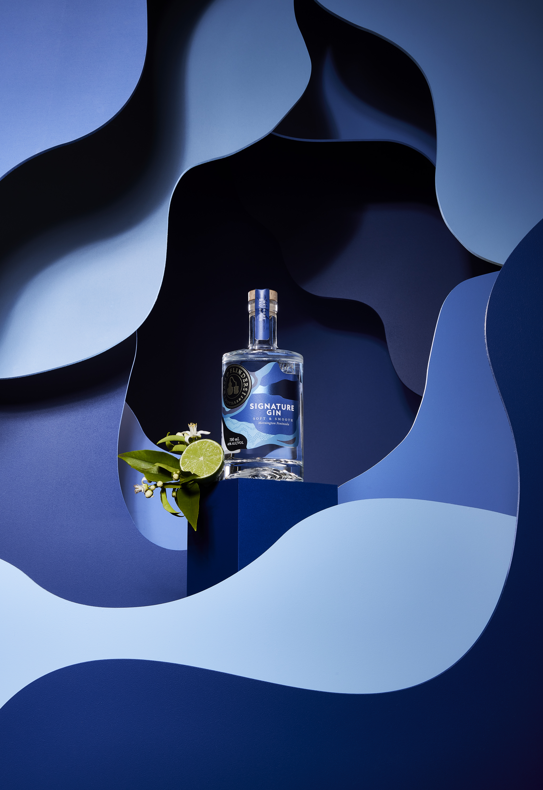 A bottle of gin sits on a blue cube with lime and flowers, against a layered blue paper cutout backdrop, showcasing a refreshing drink.