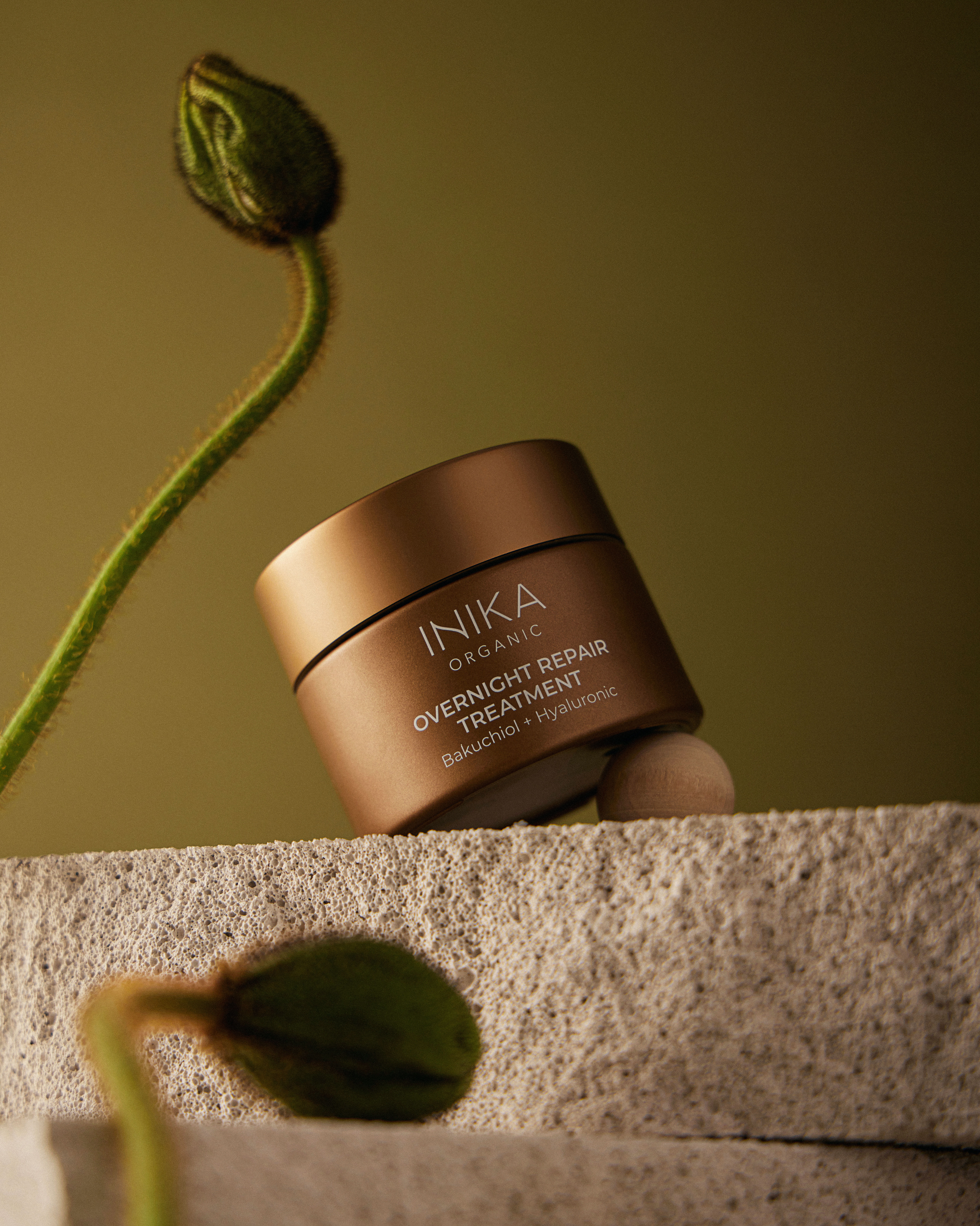Inika Organic overnight repair treatment jar on a concrete platform with flower buds against a green background in a beauty product shot.