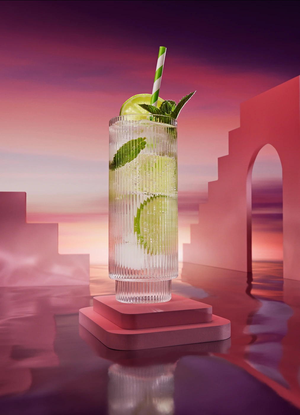 Tall ribbed glass cocktail with lime slices, mint leaves, and striped straw on a pink platform against sunset sky
