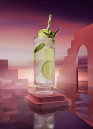 Tall ribbed glass cocktail with lime slices, mint leaves, and striped straw on a pink platform against sunset sky