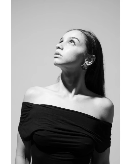 Black and white portrait of a young adult woman wearing off-the-shoulder top and earrings, looking up against a plain background.