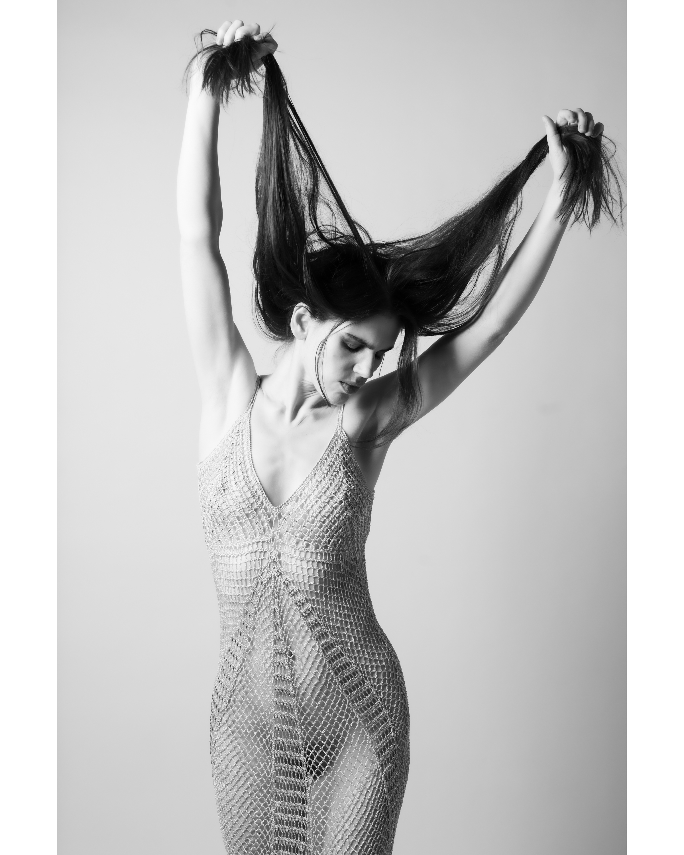 Black and white portrait of a female adult model in an open knit dress, holding her long hair above her head in a studio setting.