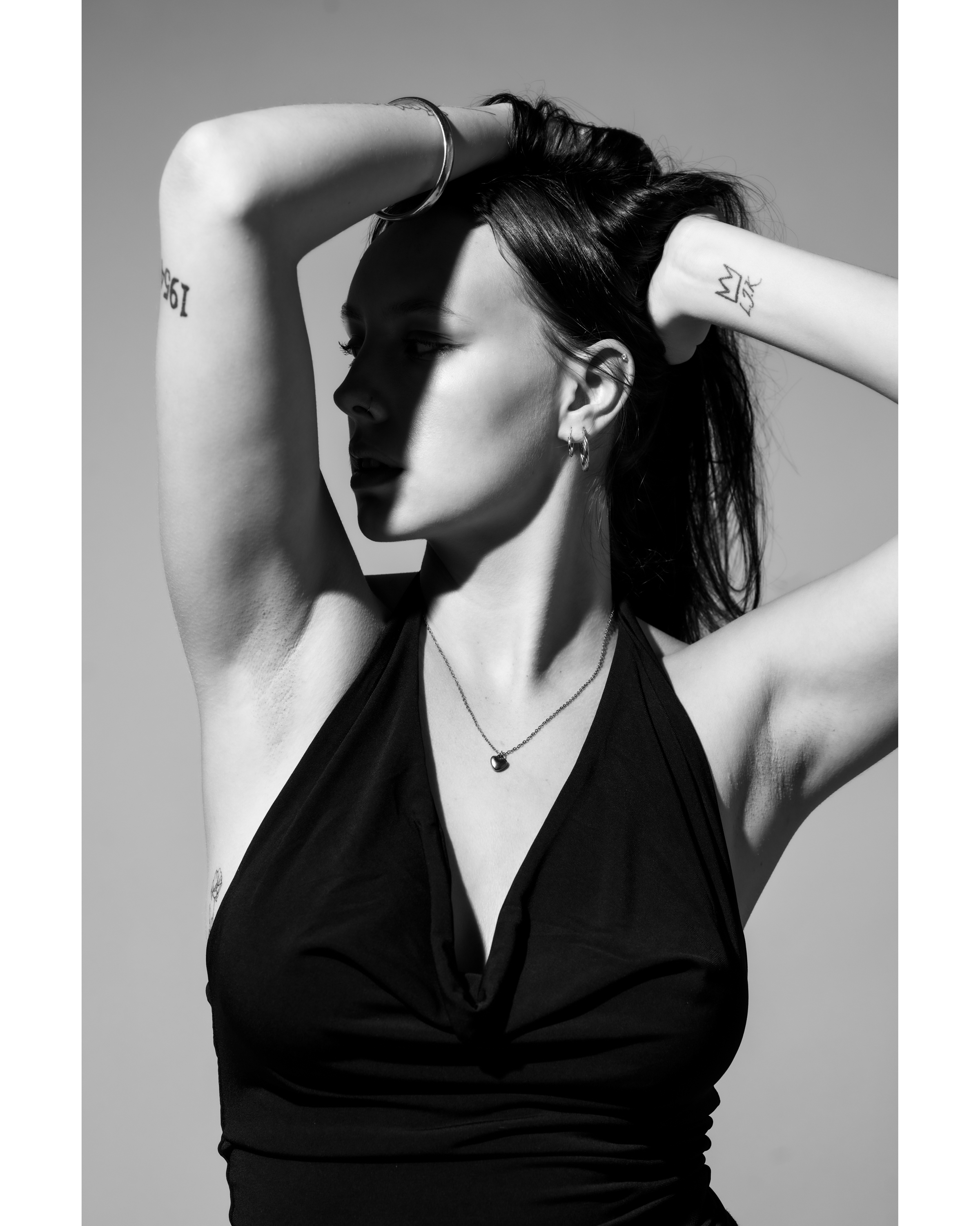 Monochrome portrait of a woman in a black halter top with her arms raised, posing against a light background.