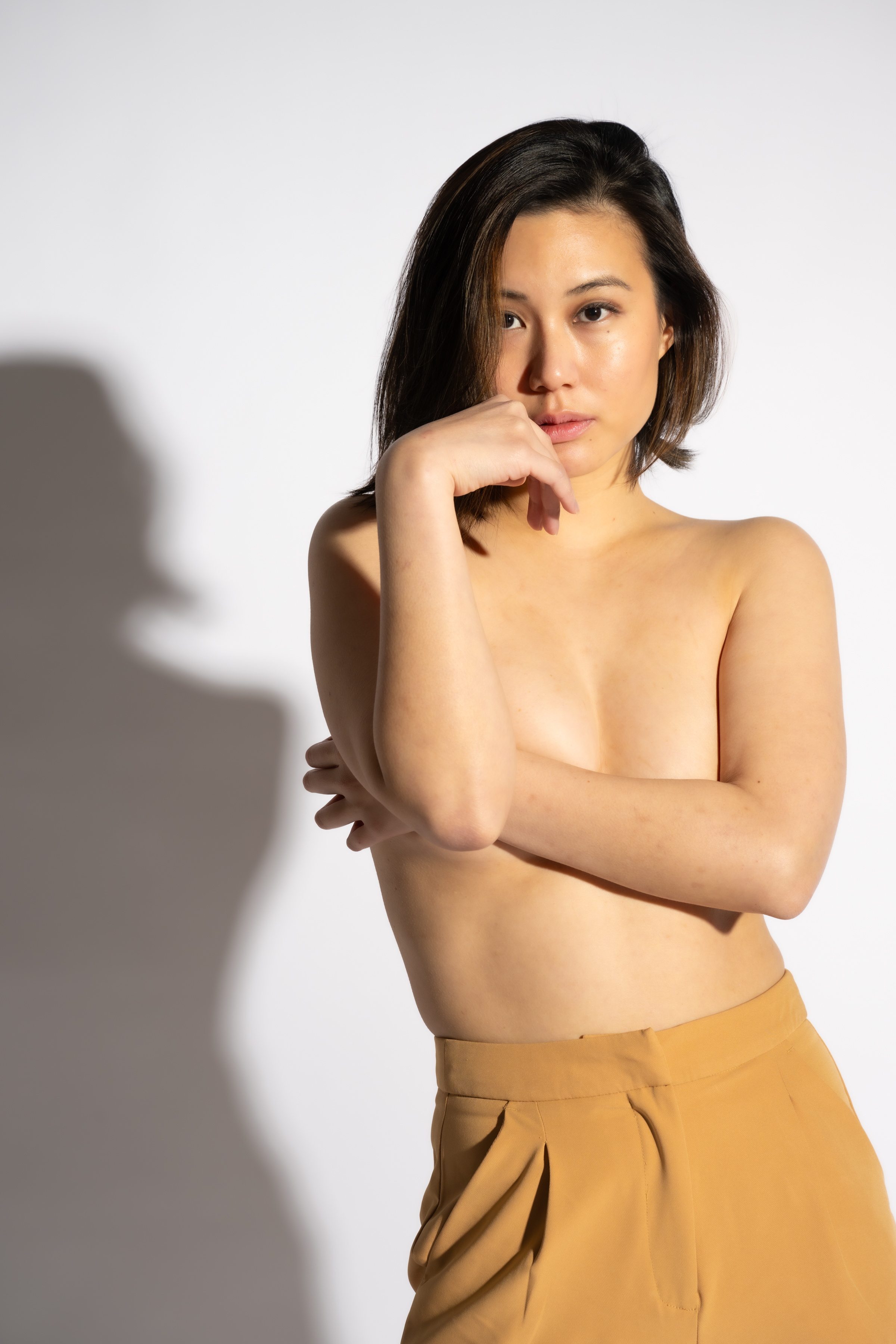 Portrait of a young adult woman with tanned skin, wearing high-waisted tan trousers, posing in front of a bright white backdrop.