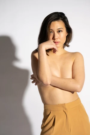 Portrait of a young adult woman with tanned skin, wearing high-waisted tan trousers, posing in front of a bright white backdrop.