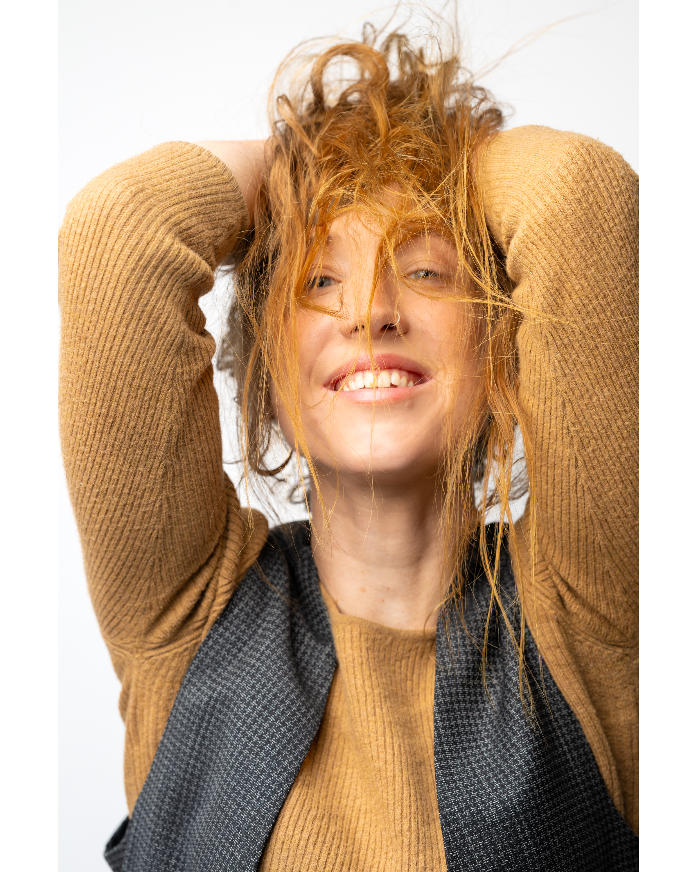 Smiling woman with wild red hair pulling at her strands, wearing a mustard knit sweater and grey patterned vest against a plain white background.