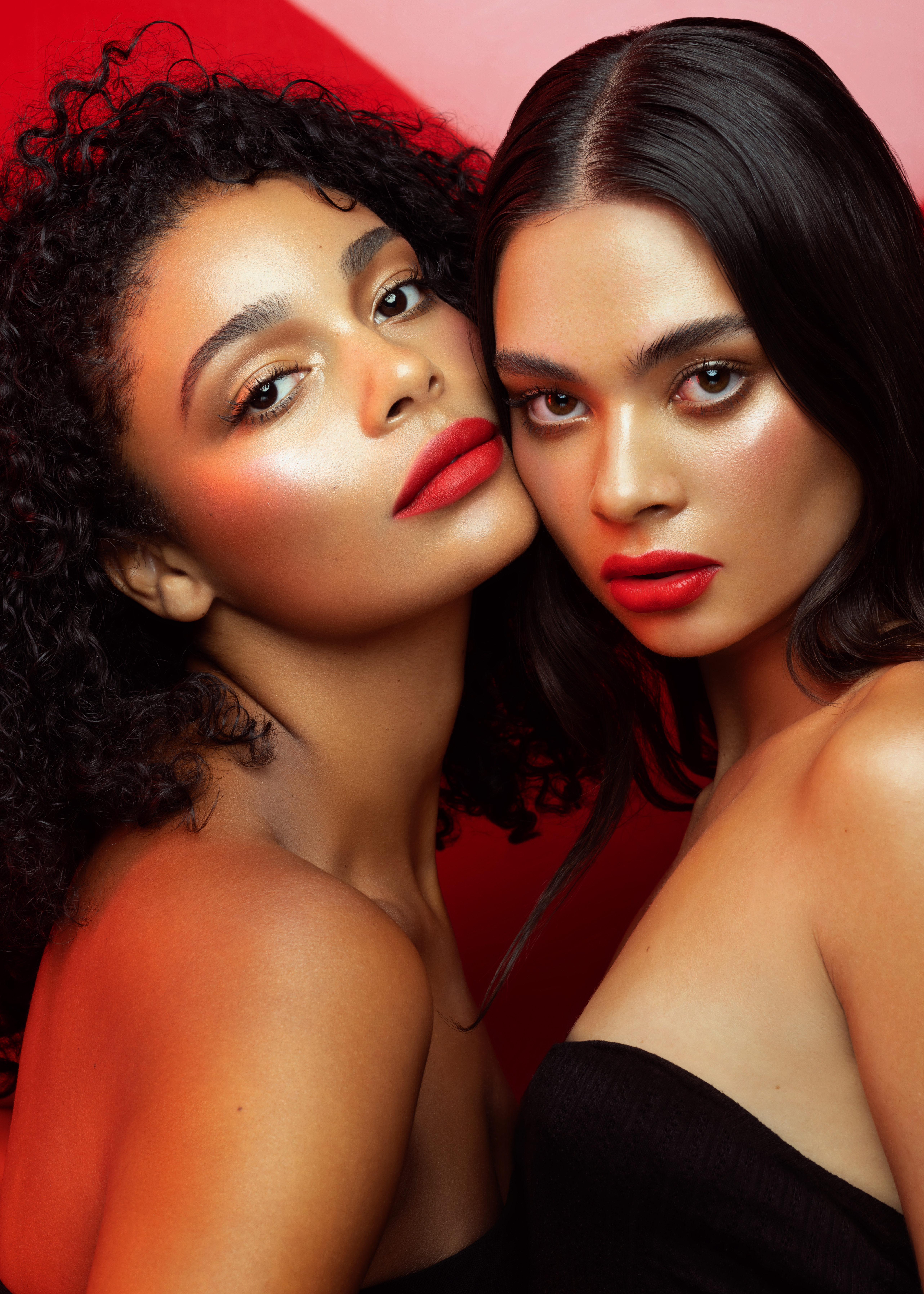 Close-up portrait of two adult women with red lips, minimal black dresses, and radiant skin posing in front of a vibrant red backdrop.