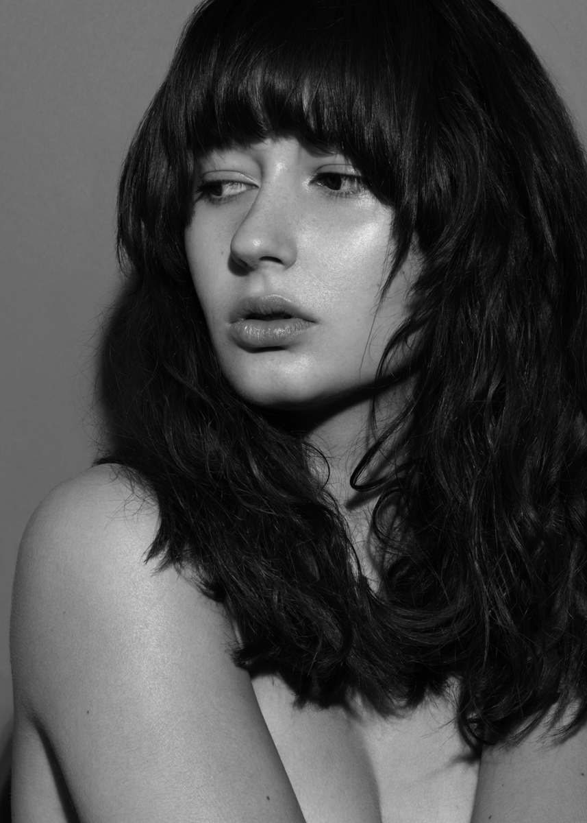 Monochrome portrait of an adult woman with long wavy dark hair and bangs, looking left, against a neutral-toned background in a studio setting.