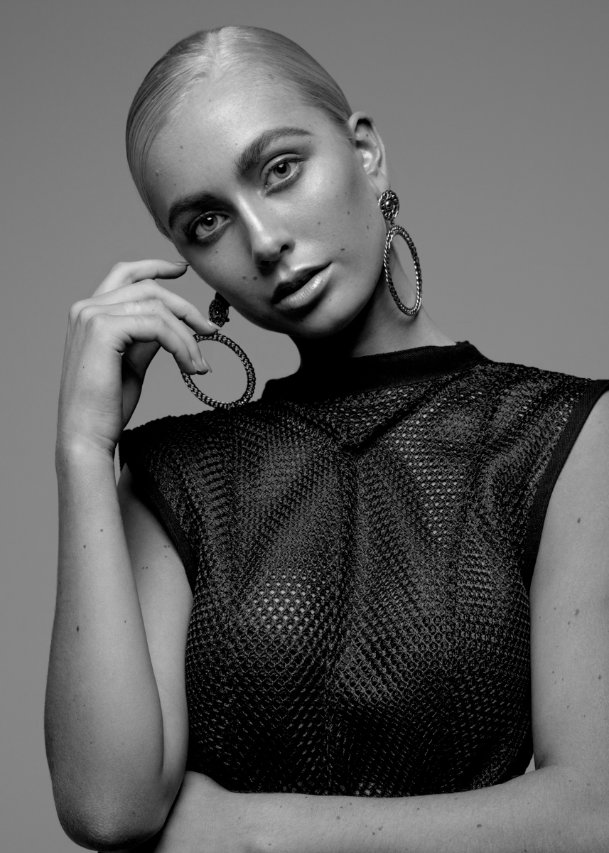 Black and white portrait of a blonde woman with an intense gaze, featuring hoop earrings and a see-through, patterned top against a grey backdrop.