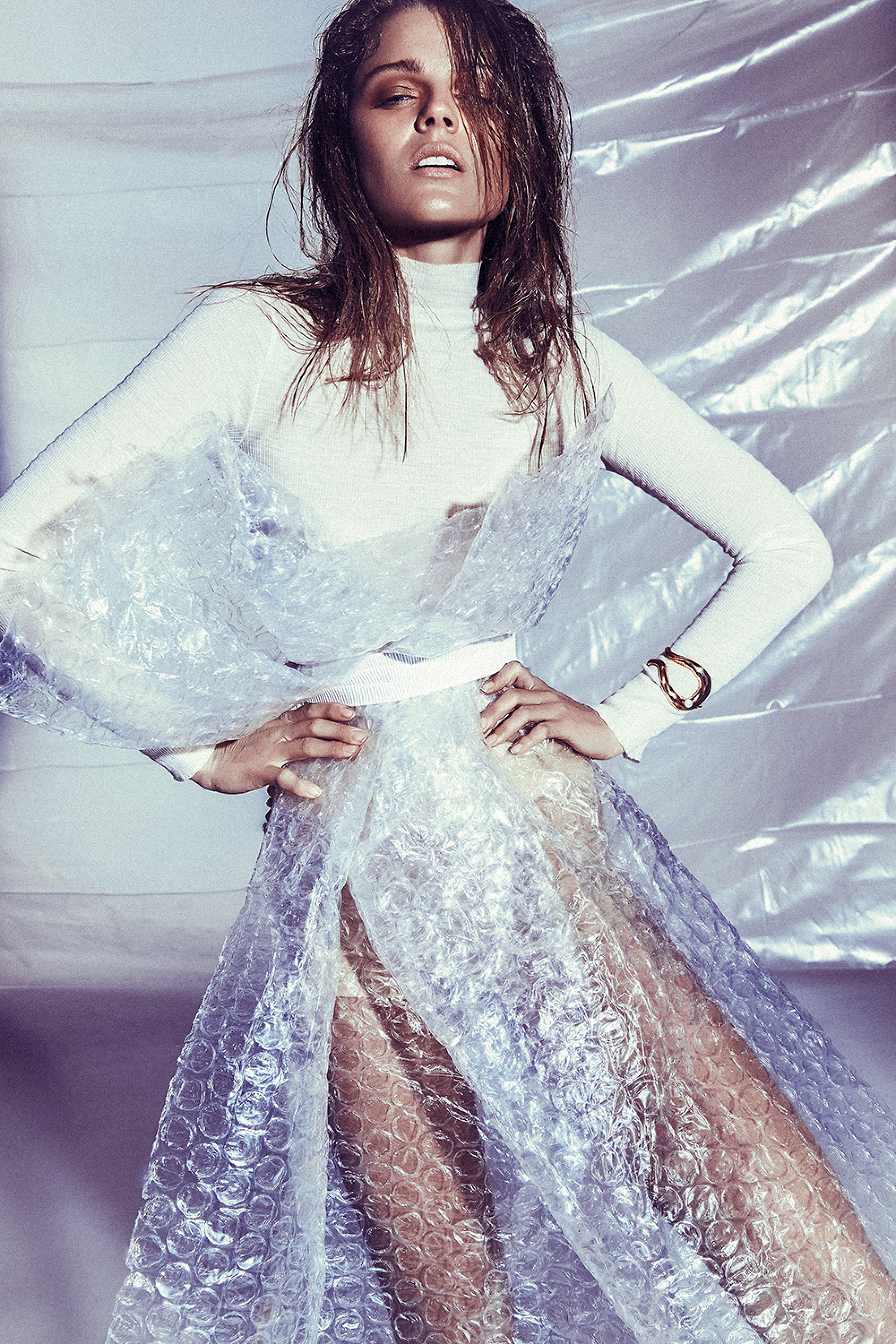 Woman in a clear bubble wrap dress with a white turtleneck poses in a studio with a wet hair look and bronze bracelet. Fashion shot.