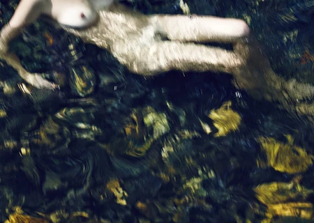 Abstract view of a nude person submerged in water over rocks, creating a blurred and dreamy effect with reflections and refractions.