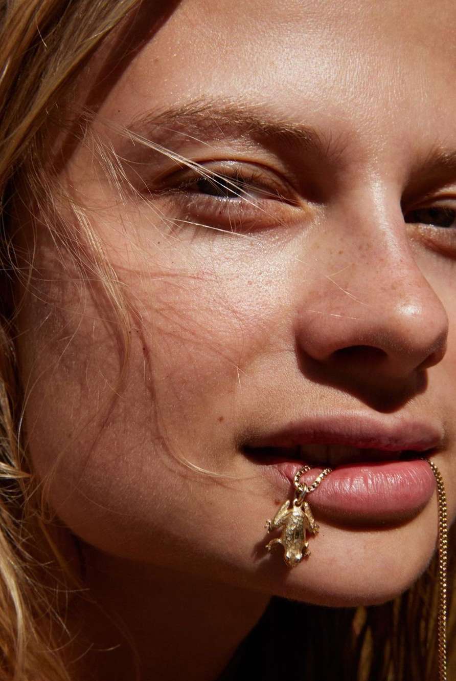 Close-up studio shot of a blonde adult woman with a gold frog pendant necklace entering her mouth. Beauty and fashion concept.