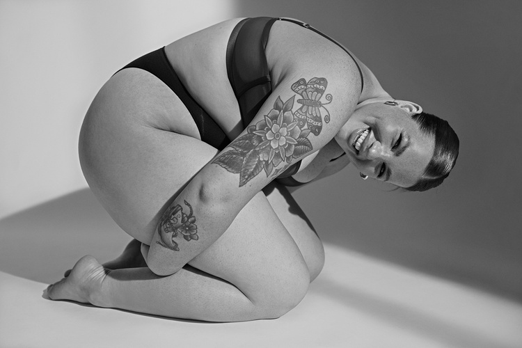Plus size woman in underwear kneeling and smiling, featuring floral tattoos on her arm, shot in monochrome.