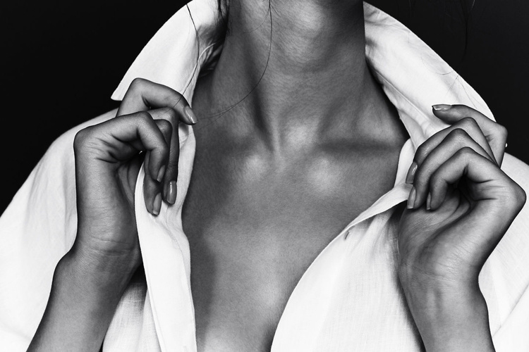 Elegant studio shot of a woman's upper body in a white collared shirt, emphasizing the collarbone and hands against a dark backdrop.