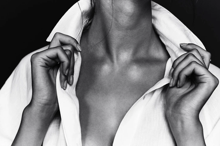 Elegant studio shot of a woman's upper body in a white collared shirt, emphasizing the collarbone and hands against a dark backdrop.