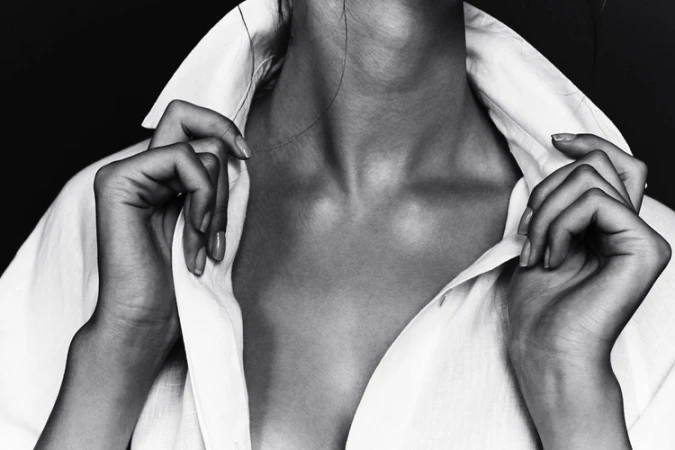 Elegant studio shot of a woman's upper body in a white collared shirt, emphasizing the collarbone and hands against a dark backdrop.