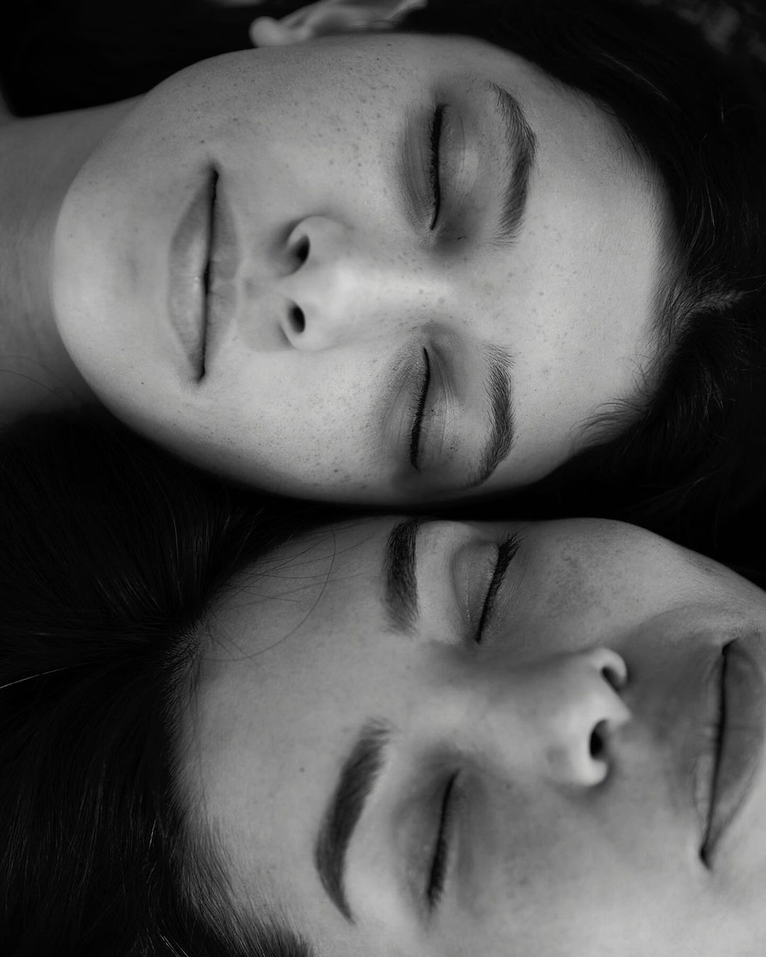 Black and white portrait of two adult women with closed eyes, resting their heads together, showcasing a serene and intimate moment.