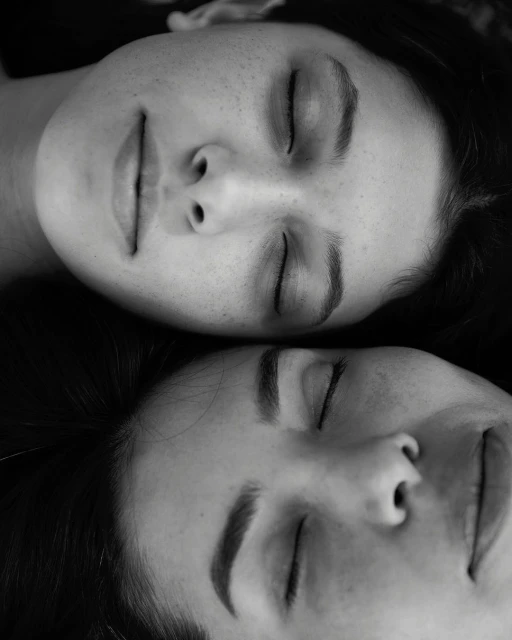 Black and white portrait of two adult women with closed eyes, resting their heads together, showcasing a serene and intimate moment.