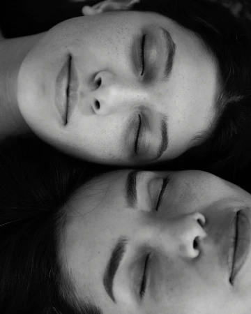 Black and white portrait of two adult women with closed eyes, resting their heads together, showcasing a serene and intimate moment.