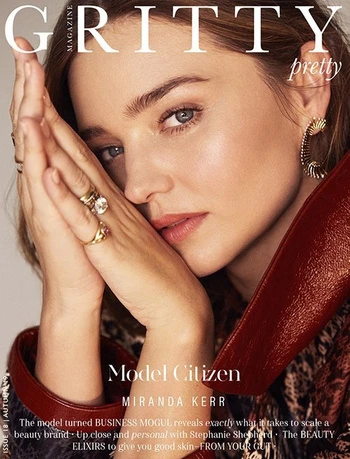 Miranda Kerr graces a magazine cover, her hands elegantly framing her face adorned with rings, makeup, and earrings under soft lighting.
