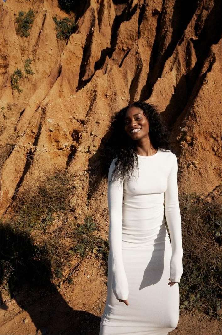 A Black woman smiles, wearing a white long-sleeve dress, posed against a striking background of reddish-brown rock formations.