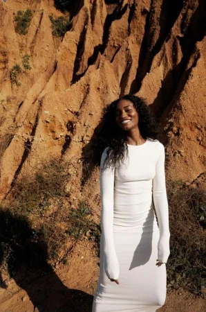 A Black woman smiles, wearing a white long-sleeve dress, posed against a striking background of reddish-brown rock formations.