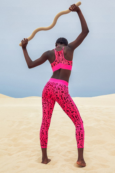 Athletic woman in hot pink activewear balances a curved wooden bar on a tan sand dune under a pale blue sky, showcasing sport and fitness.