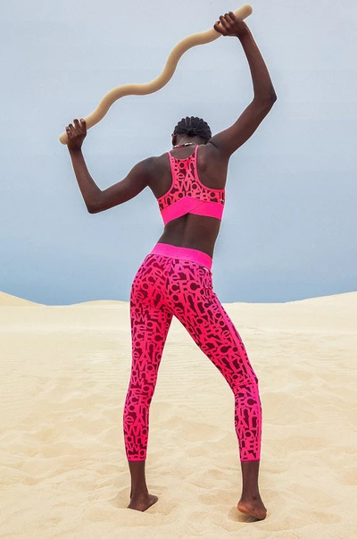 Athletic woman in hot pink activewear balances a curved wooden bar on a tan sand dune under a pale blue sky, showcasing sport and fitness.