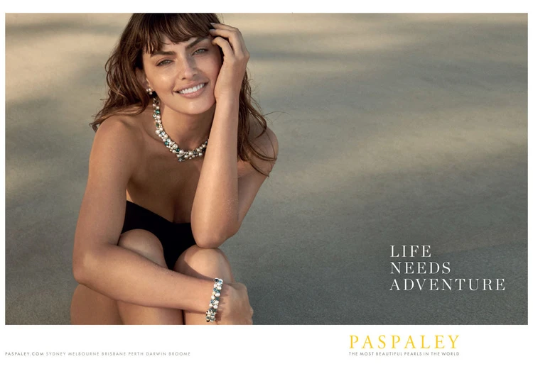 Brunette woman smiles on beach wearing pearl necklace and bracelet. Close-up fashion shot in soft, neutral light with tan sand backdrop.