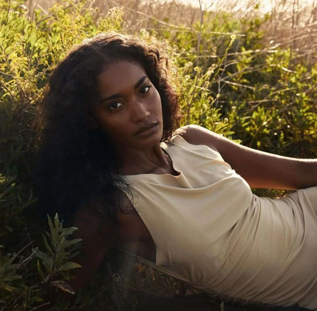 Elegant woman with long curly hair in a cream dress lies in a sunlit meadow. Fashion shot in natural light in an outdoor setting.